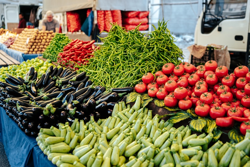 A market stall displays piles of fresh vegetables, including tomatoes, green peppers, eggplants, zucchinis, and red chili peppers, with bags of produce and a vendor in the background.