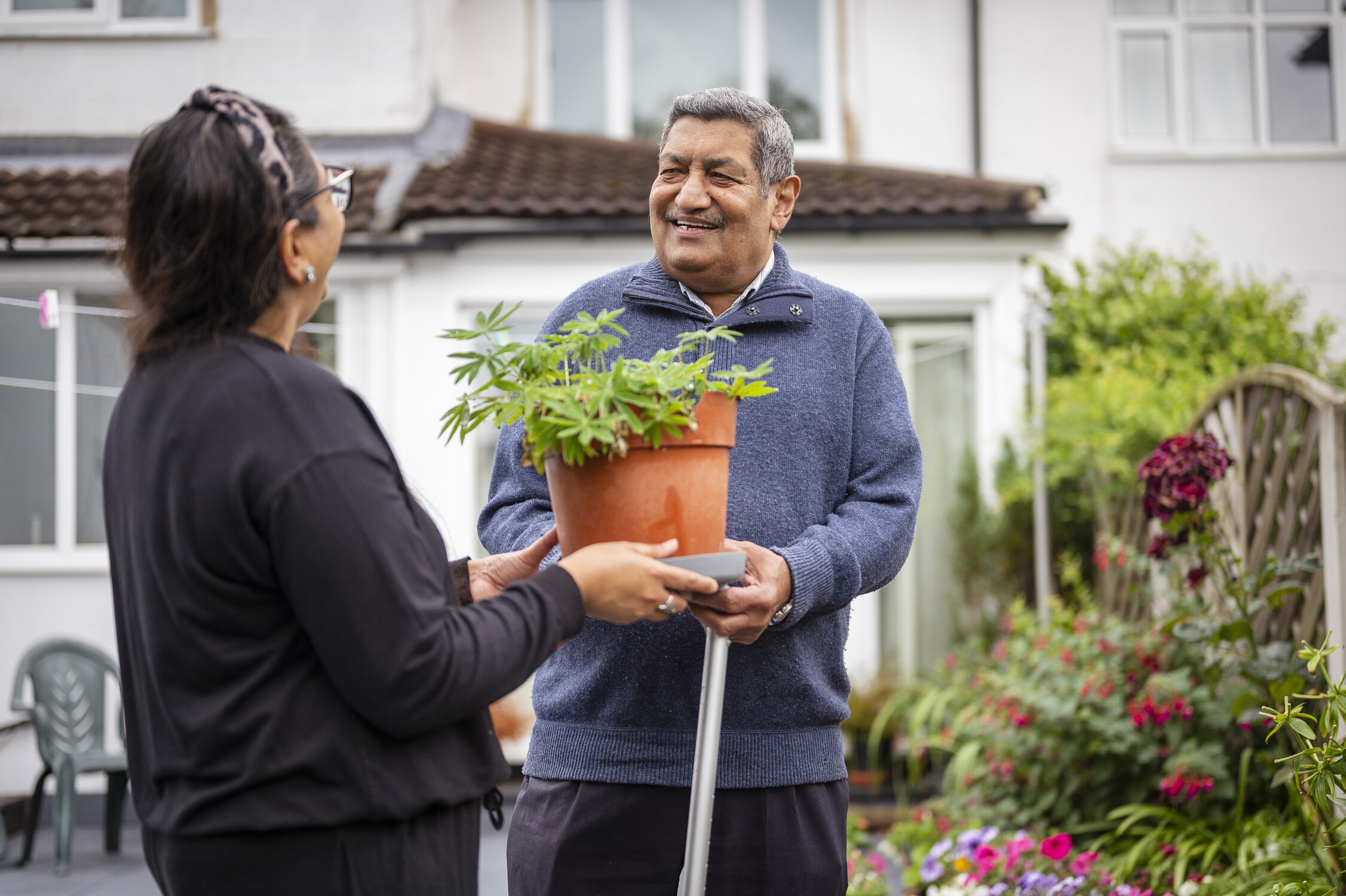 Two people stand in a garden. One hands a potted plant to the other. A house with a tiled roof and flowers is in the background. Their faces are blurred.