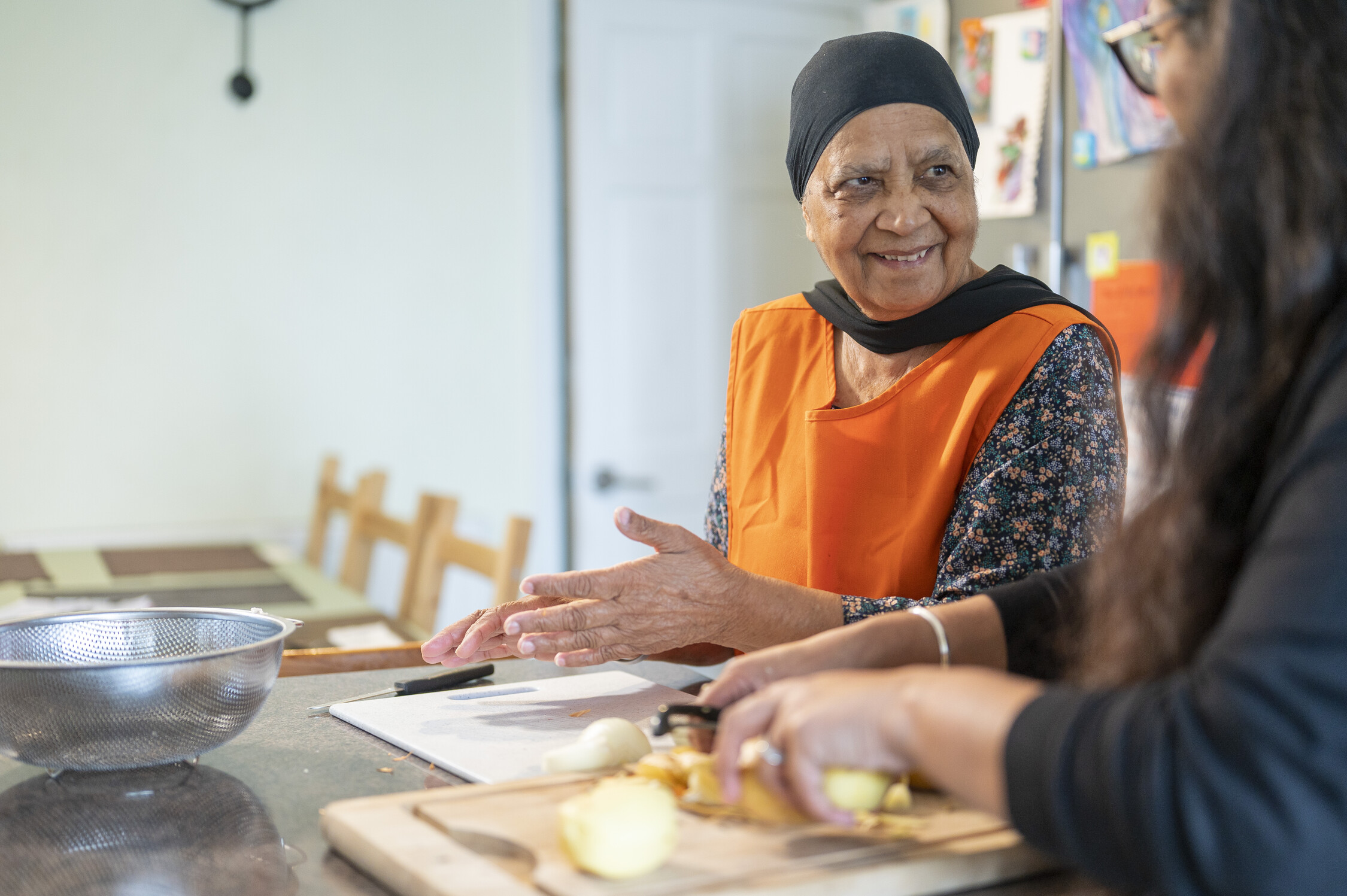 Cooking Two people in a kitchen, one seated with clasped hands wearing an orange apron and headscarf, the other chopping onions near a colander.