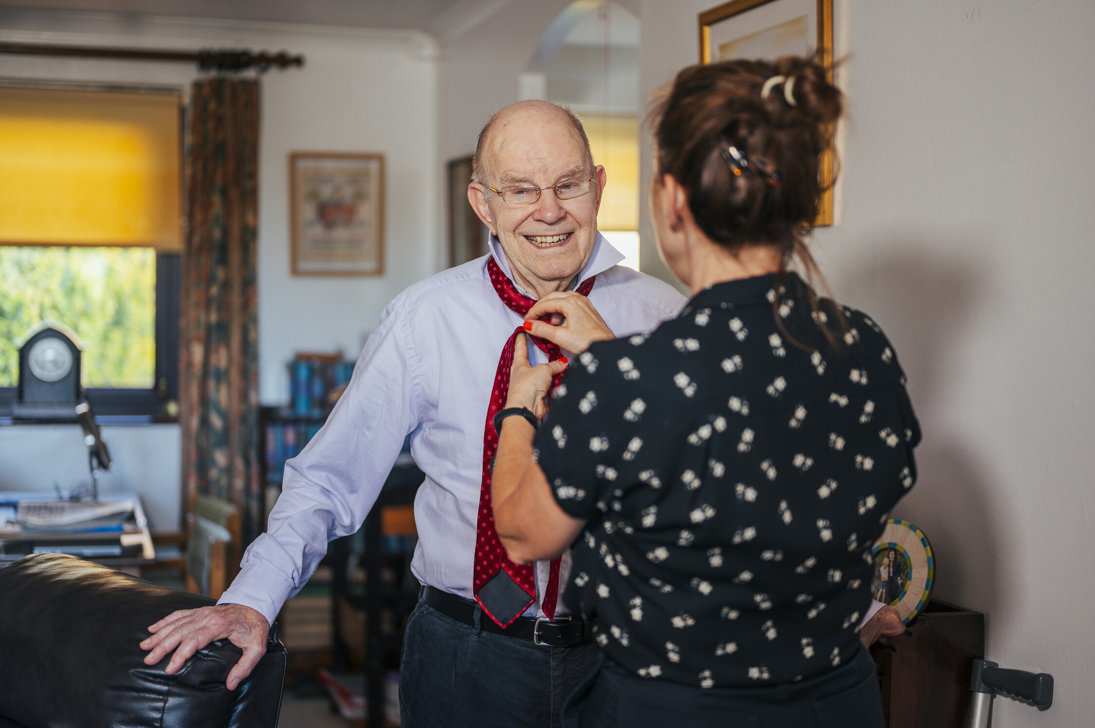 A woman helps a man tie a red and black necktie. He wears a light purple shirt and dark trousers. She wears a black blouse with white patterns. They are in a bright room with a leather chair and bookshelves,