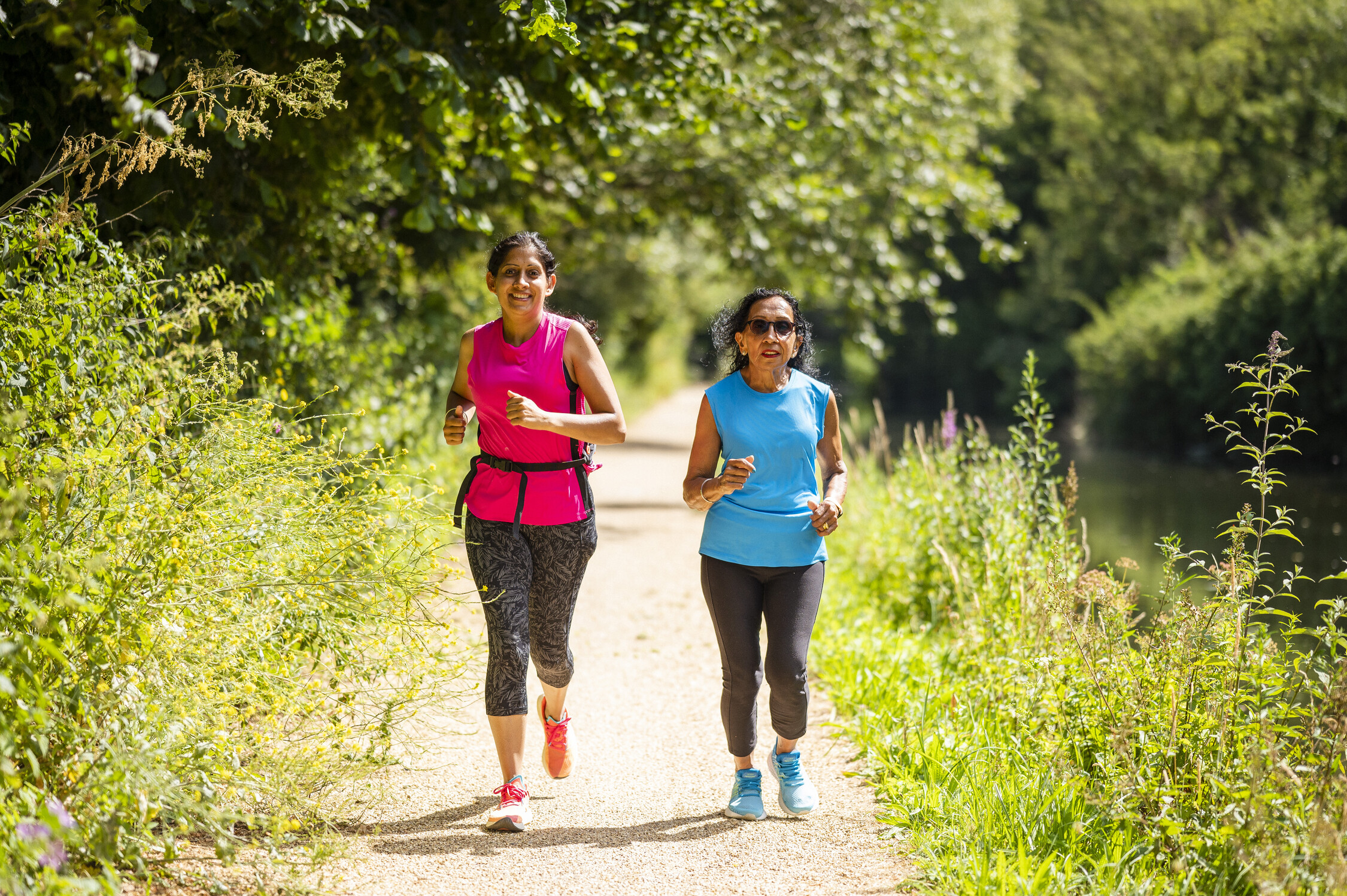 Two women jog on a path with trees and tall grass. One wears a pink top, the other a blue top. Both wear black leggings.