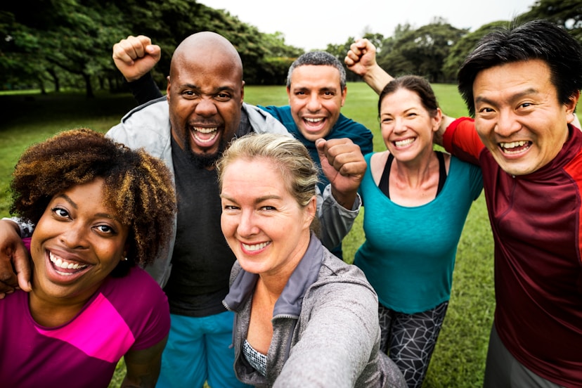 A diverse group of six adults smile and cheer together outdoors, posing for a selfie in a grassy park, expressing happiness and camaraderie.