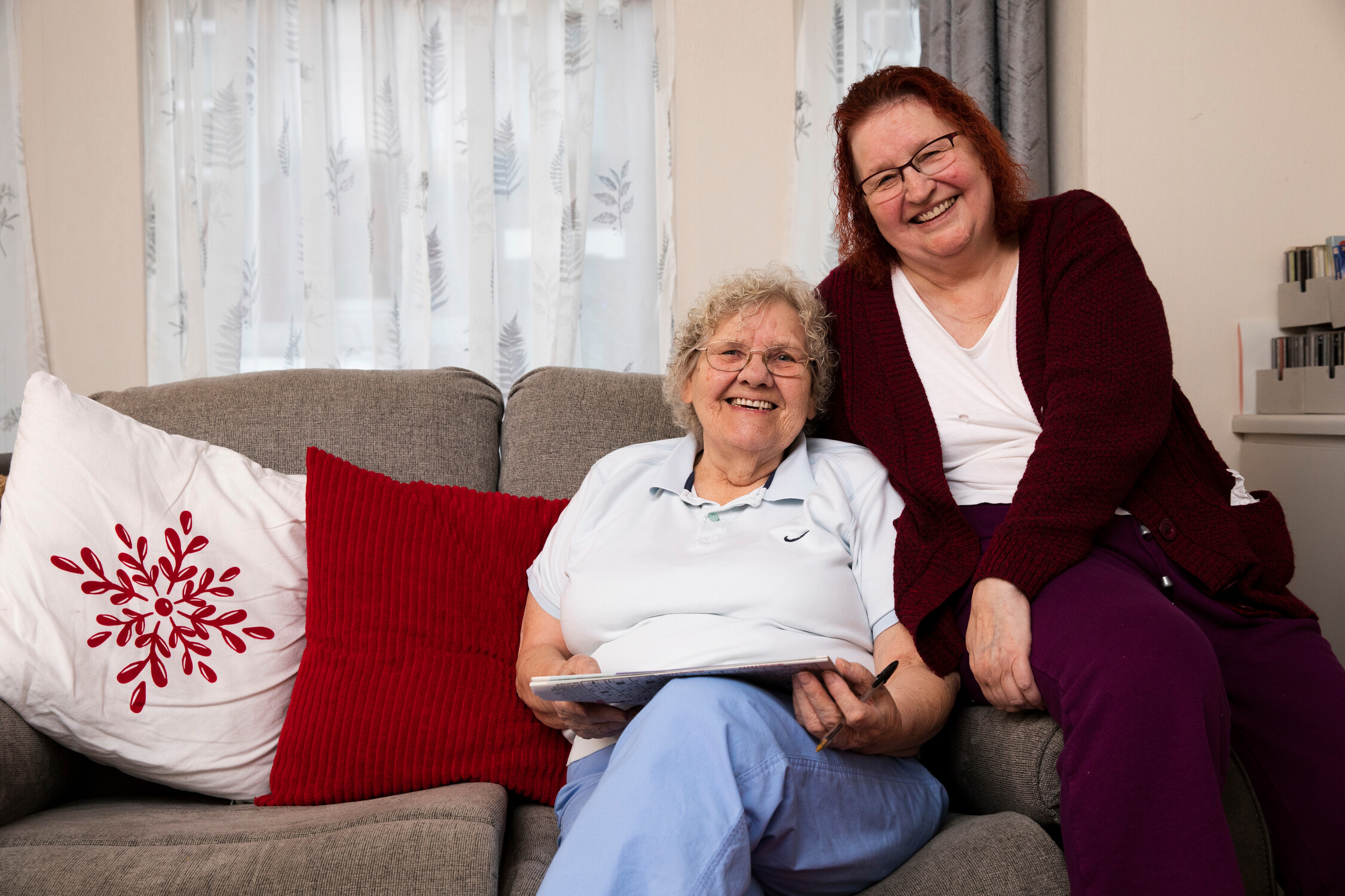 Two people on a gray sofa, one holding a tablet, with red pillows and leaf-patterned curtains in the background.