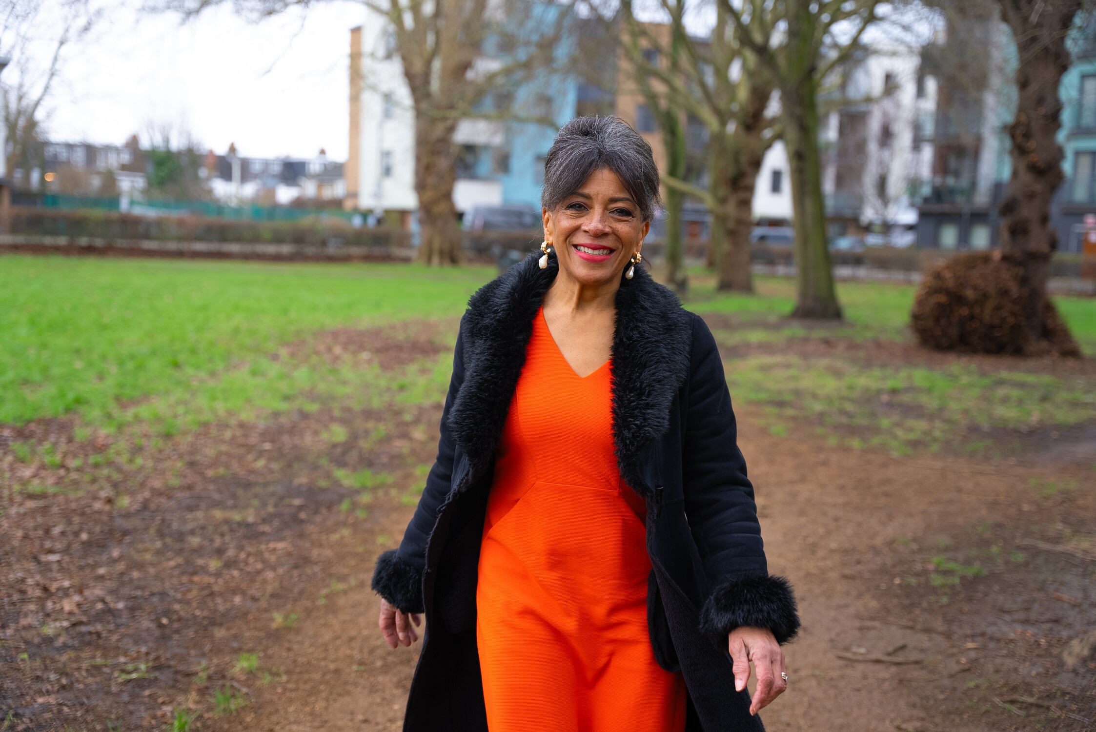 A person walks on a dirt path in a park. They wear an orange outfit and a black coat with fur trim. Grass, trees, and buildings are in the background.