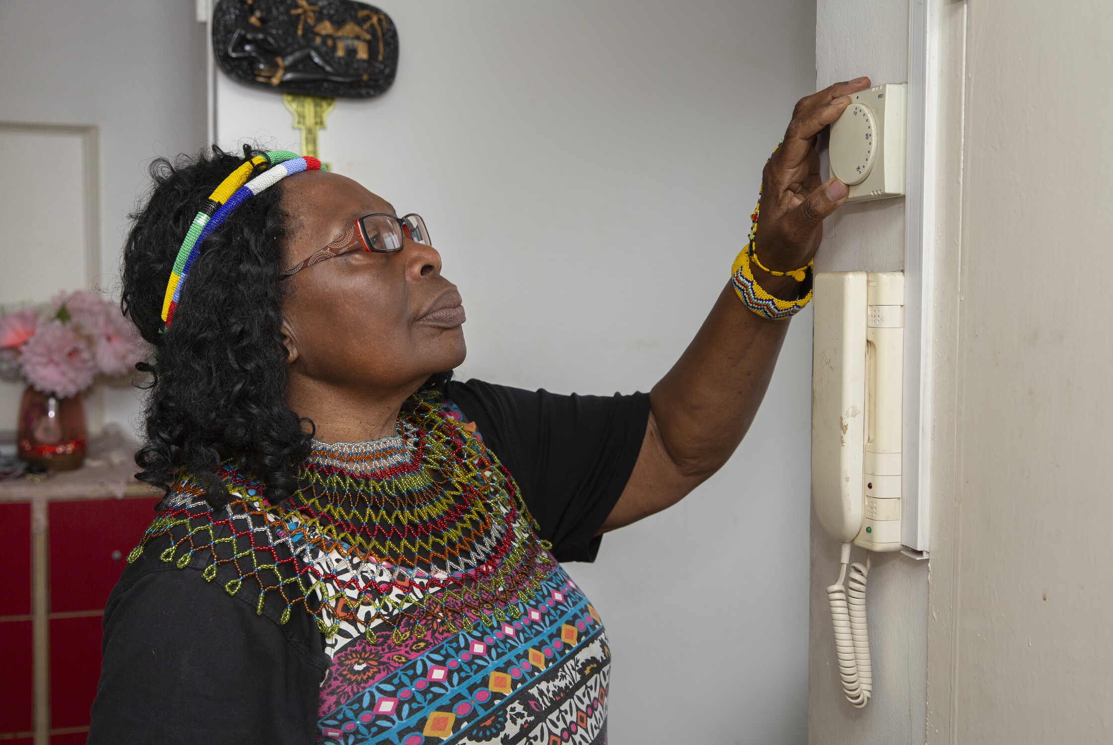 Woman adjusting a thermostat on the wall, wearing a colorful shirt and accessories, with flowers and decor in the background.