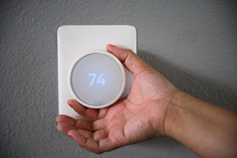 A hand adjusts a modern, round digital thermostat mounted on a gray wall. The display shows the temperature set to 74 degrees.