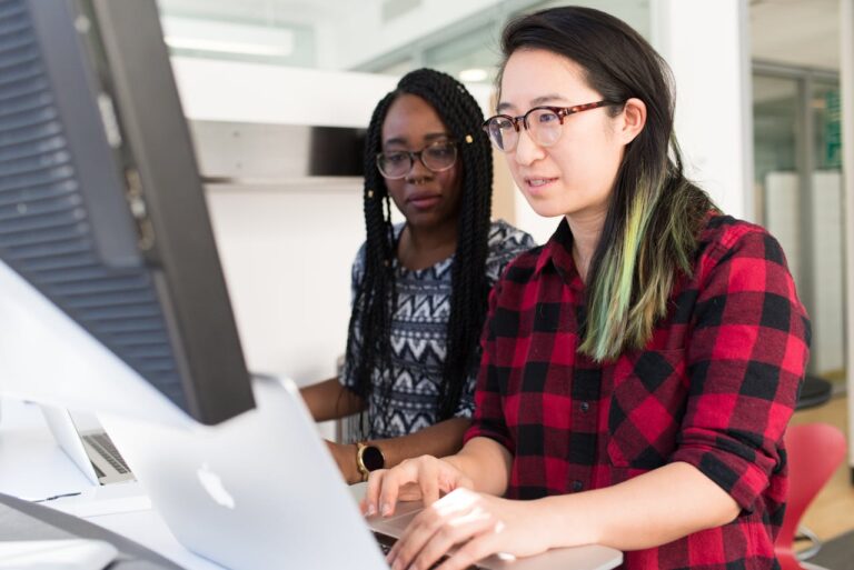 Two women sat together in front of a computer looking at the computer screen together.