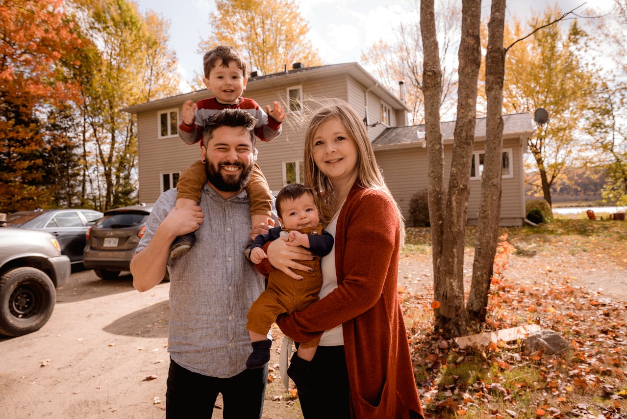 A family of four stands outside a house with autumn trees. One adult holds a child on their shoulders; the other holds an infant.