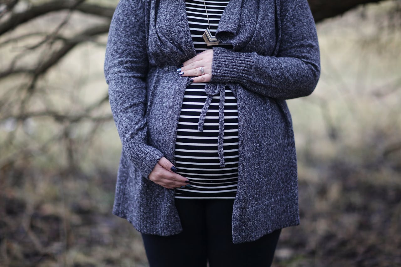 A person stands outdoors with hands on their pregnant belly. They wear a gray cardigan and striped shirt. Trees and foliage are blurred in the background