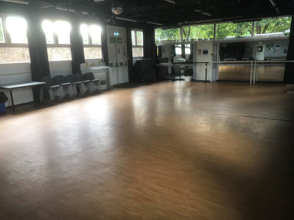 Empty room at Fairkytes with wooden floor, stacked chairs, tables, large windows, mirrored back wall, and a covered piano.