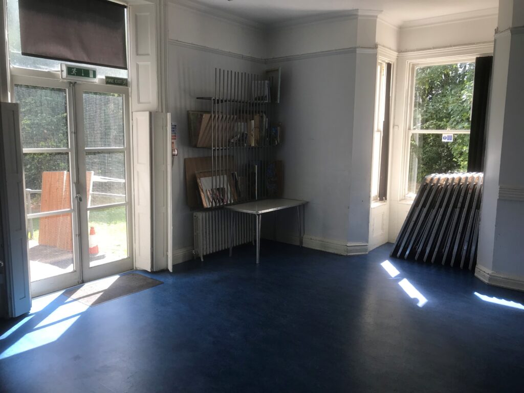 Empty room at Fairkytes Art Centre with blue flooring, white walls, double doors, a rack with boards, a small table, and stacked metal frames near a large window.