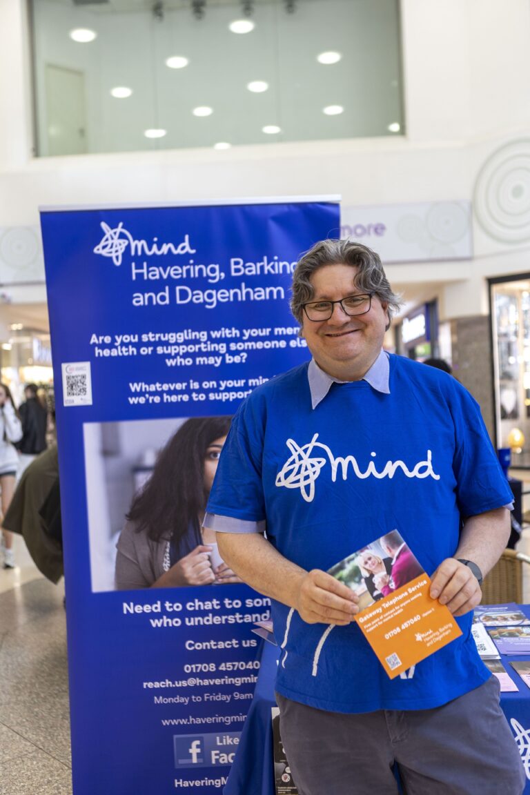 A smiling man in glasses and a blue Mind T-shirt holds a leaflet while standing in front of a Mind mental health support banner at an indoor event.