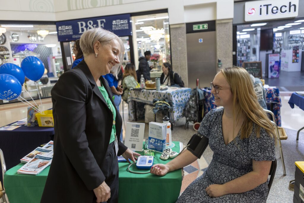A woman in a blazer smiles while checking another woman’s blood pressure at a health booth in a shopping mall. The seated woman wears a floral dress and a blood pressure cuff. Balloons and informational tables are visible in the background.