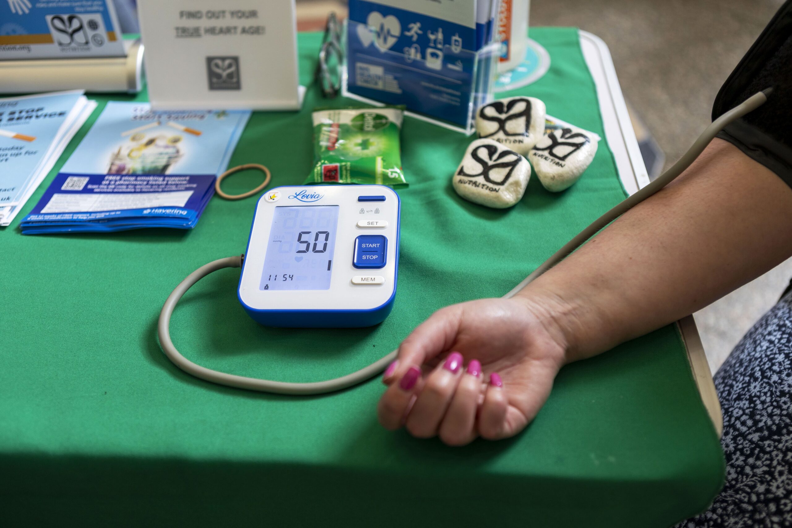 A person’s arm with painted nails is using a digital blood pressure monitor on a green table, surrounded by health brochures and promotional items. The monitor displays a reading of 50.