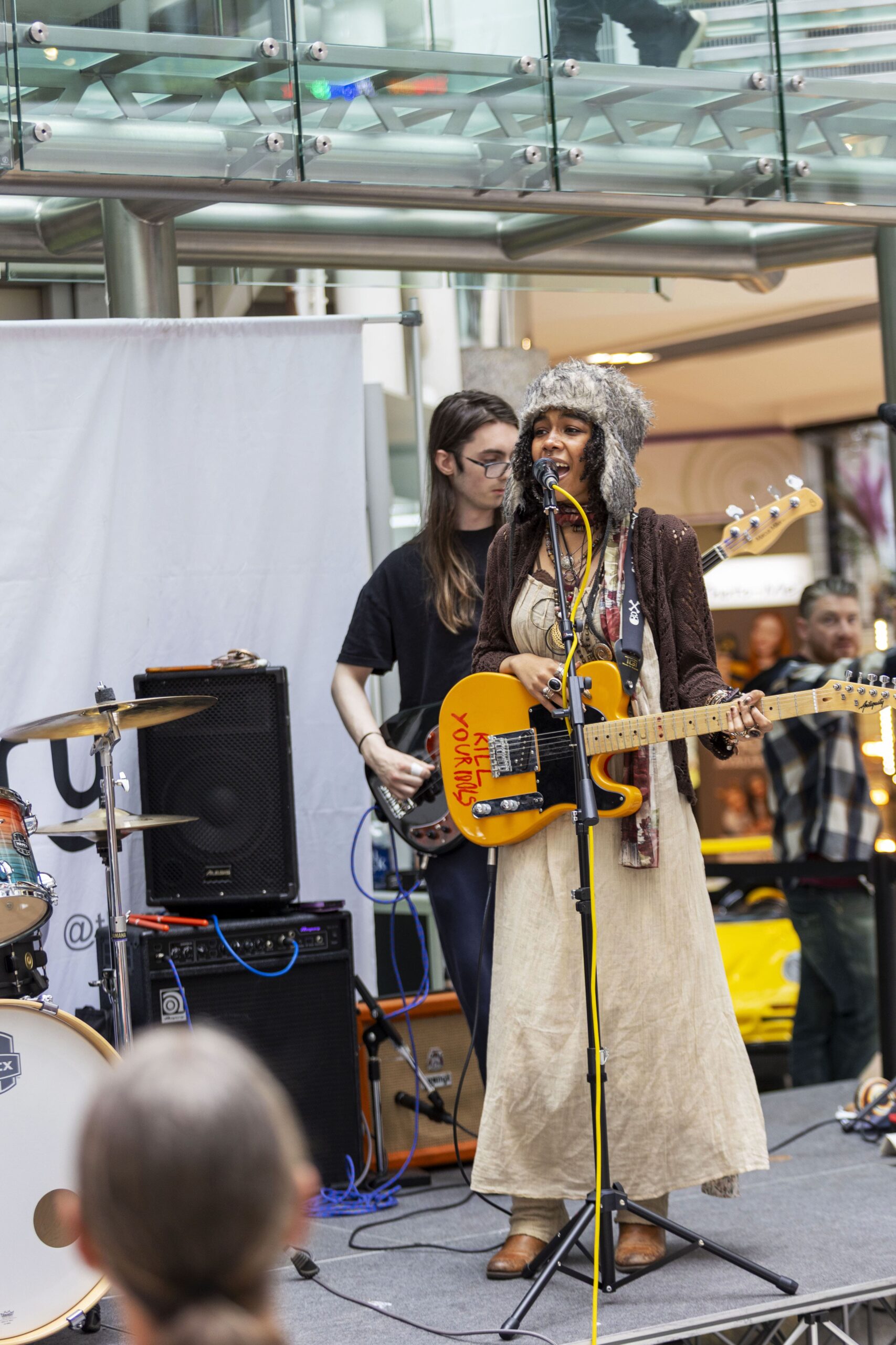 A woman in a fur hat and long dress sings and plays guitar onstage with a band. A bassist stands behind her, and a drum set is visible. They are performing in a bright indoor space with glass railings.