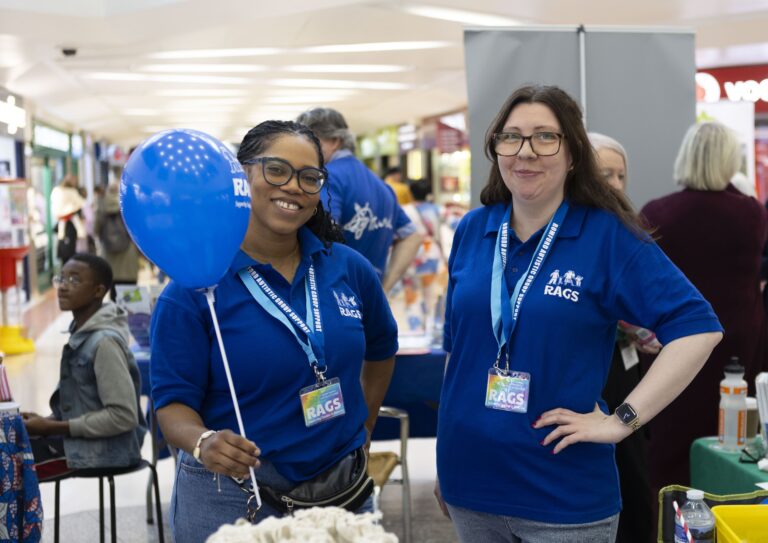 Two women wearing blue RAGS polo shirts and lanyards smile at a community event. One holds a blue balloon. They stand in a busy indoor space with tables and people in the background.