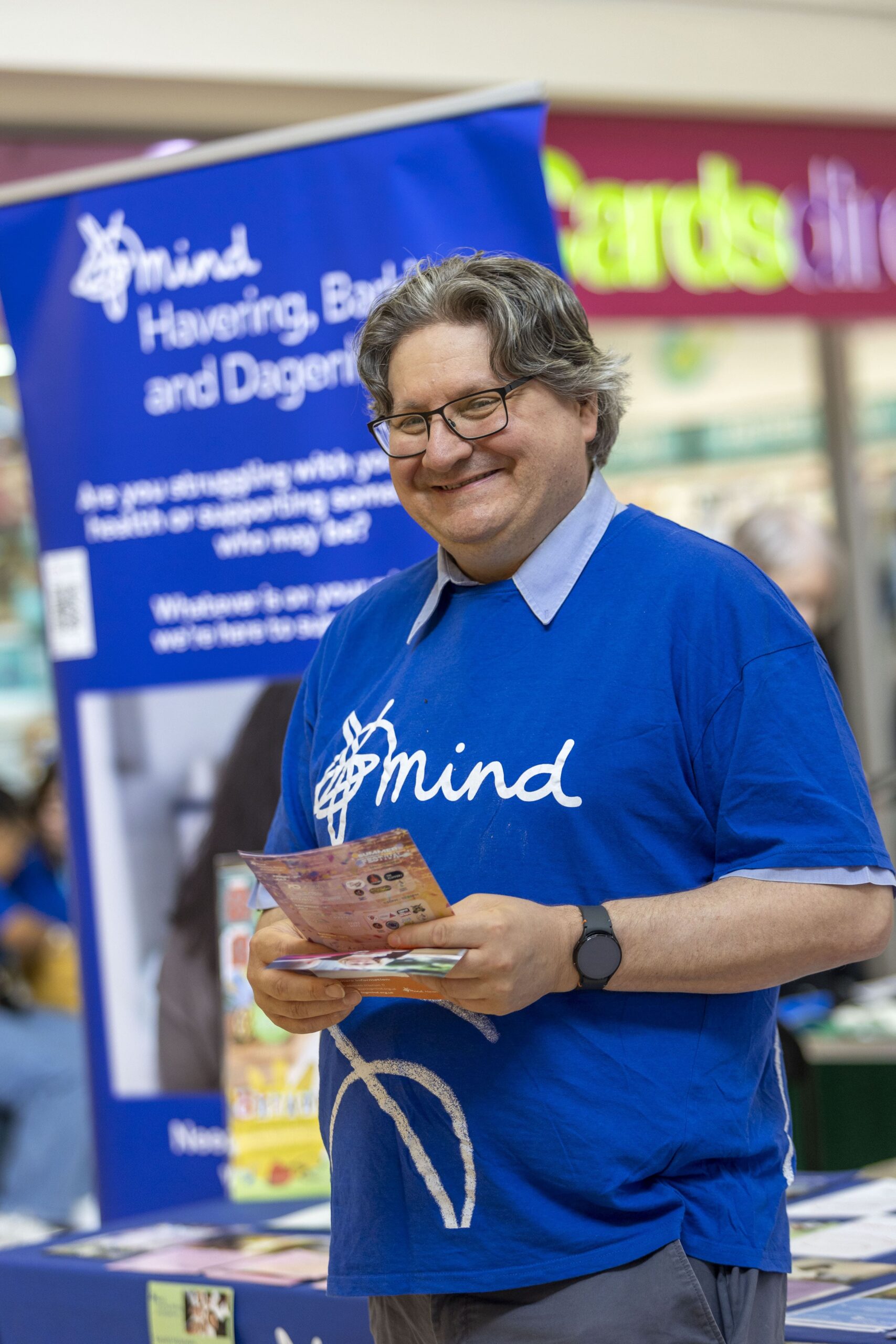 Man in a blue "Mind" t-shirt stands at a booth with a banner for Mind Havering, Barking and Dagenham, holding pamphlets.