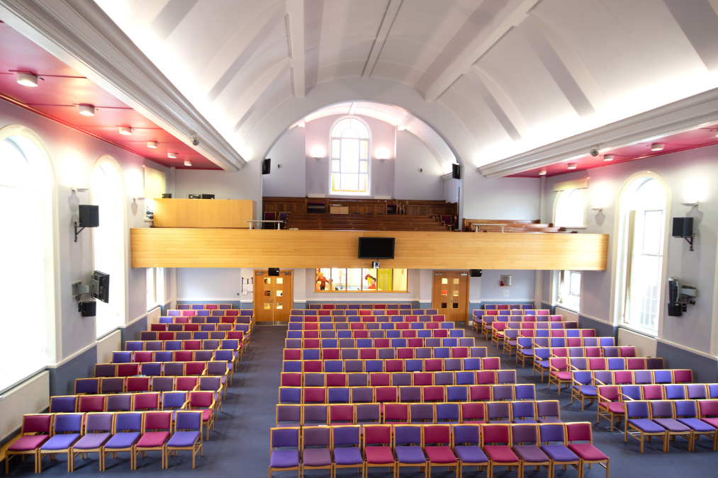 Large hall in Romford Baptist Church with rows of purple and red cushioned chairs, a balcony, arched window, and tall windows letting in natural light.