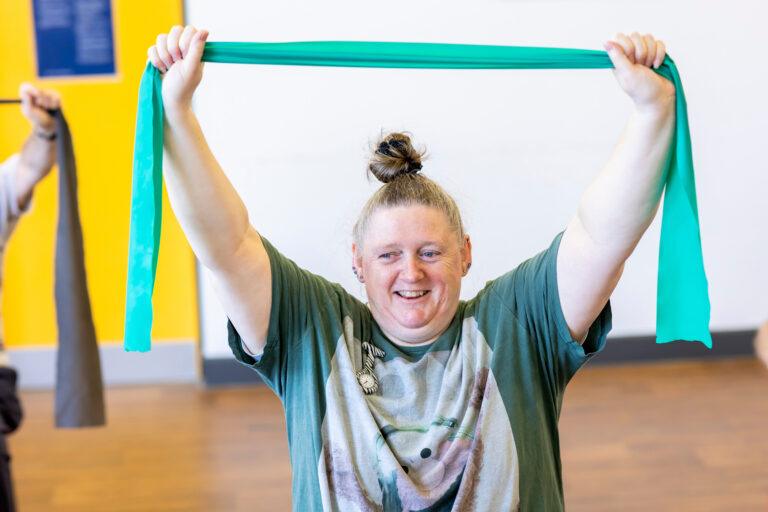 Person holding a green resistance band overhead; another person with a grey band is partially visible against a yellow wall.