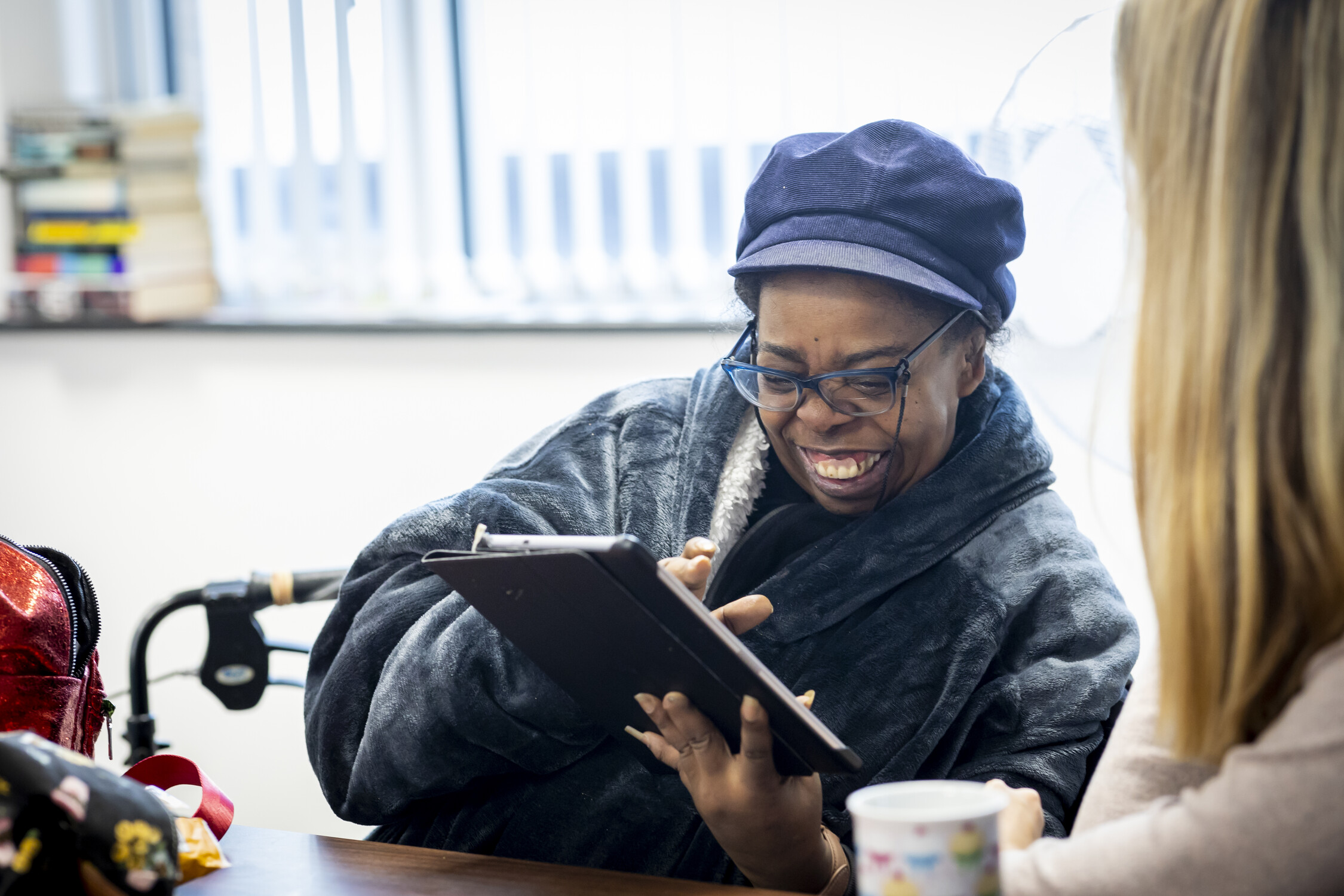 A smiling woman wearing glasses and a blue cap uses a tablet, sitting at a table with another person. Both appear engaged and happy, with books and a mug visible in the background.