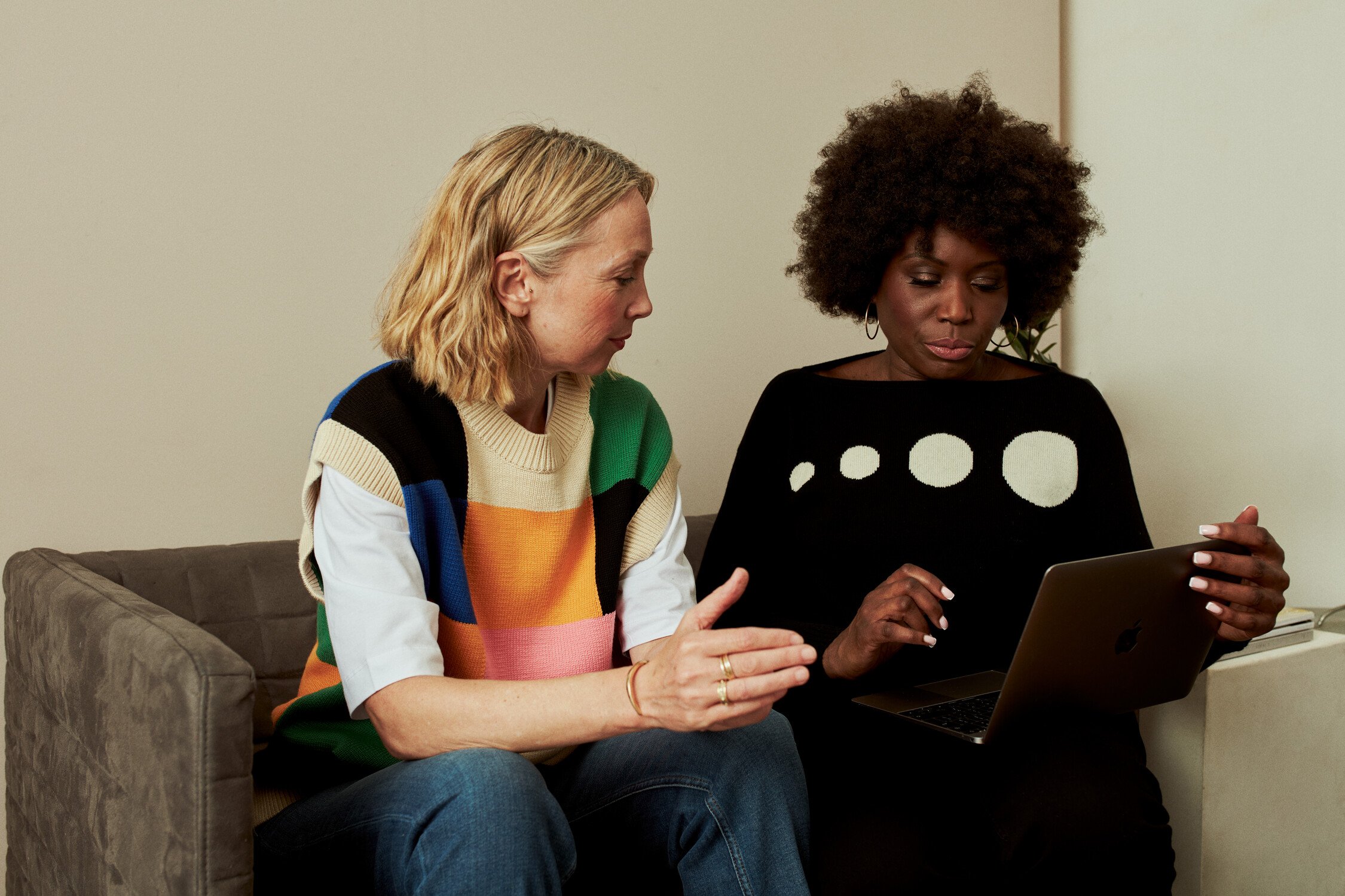 Two people sitting on a couch; one gestures while the other holds an open laptop. Faces are obscured.