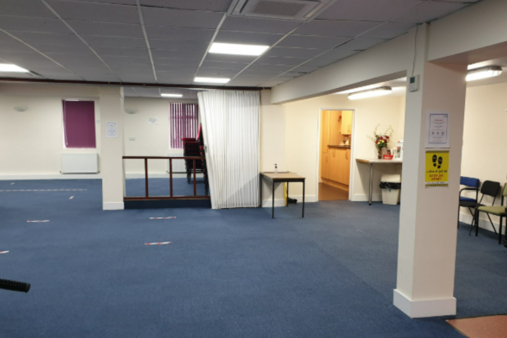 Large HASWA room with blue carpet, fluorescent lights, chairs along walls, a table near an open doorway, purple blinds, and a white curtain dividing the space.