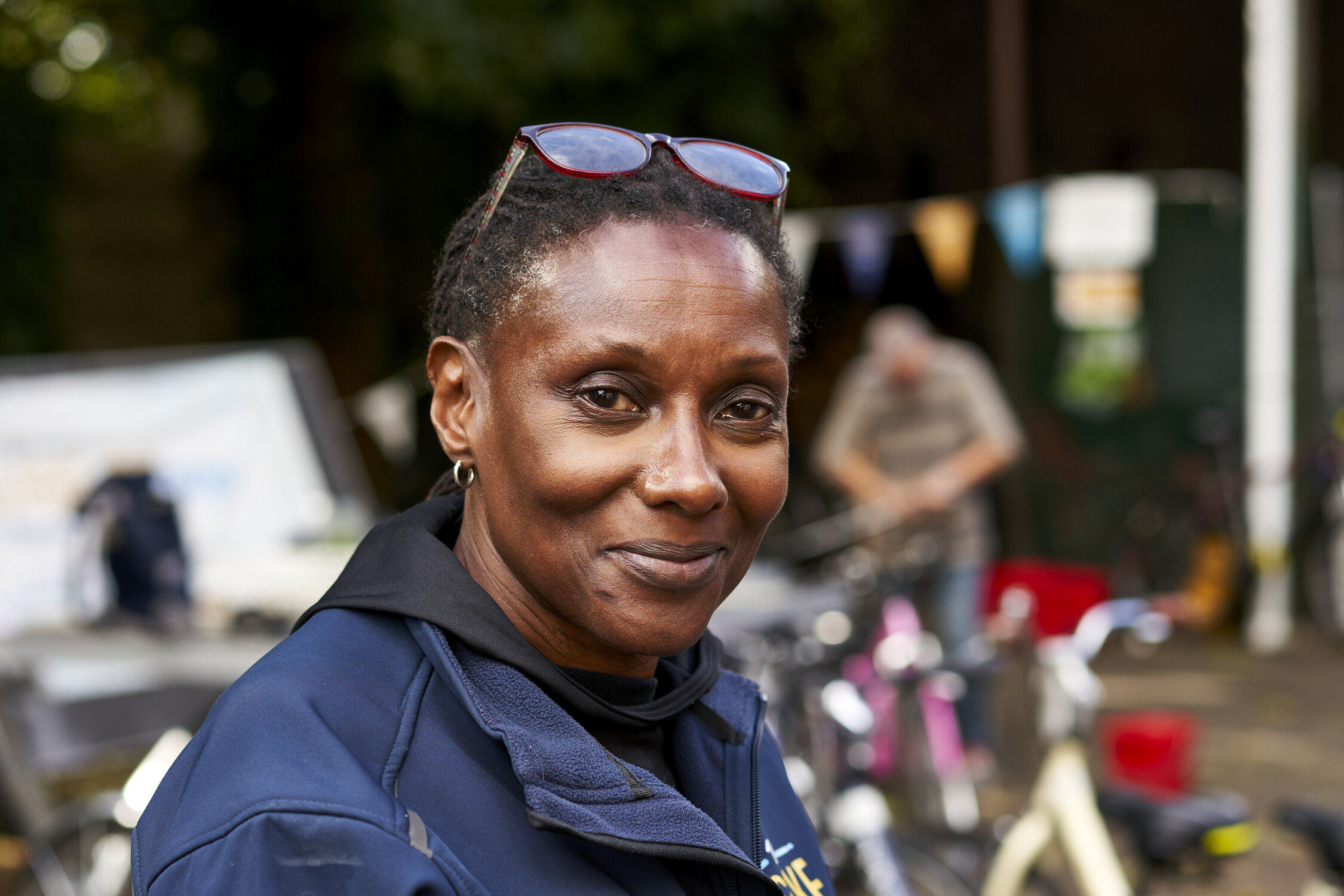A woman with short hair and glasses resting on her head smiles softly at the camera. She is wearing a navy jacket, and there are blurred bicycles and people in the background outdoors.