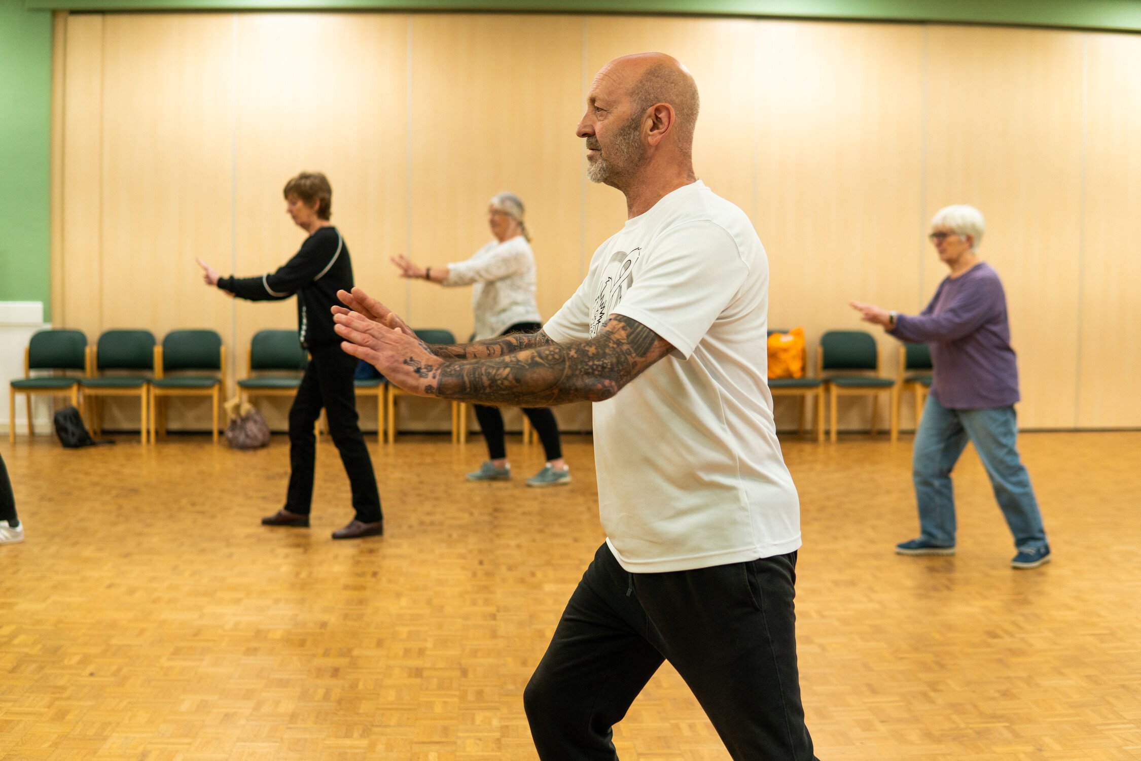 Four people practice Tai Chi in a room with wooden flooring and chairs along the back wall.