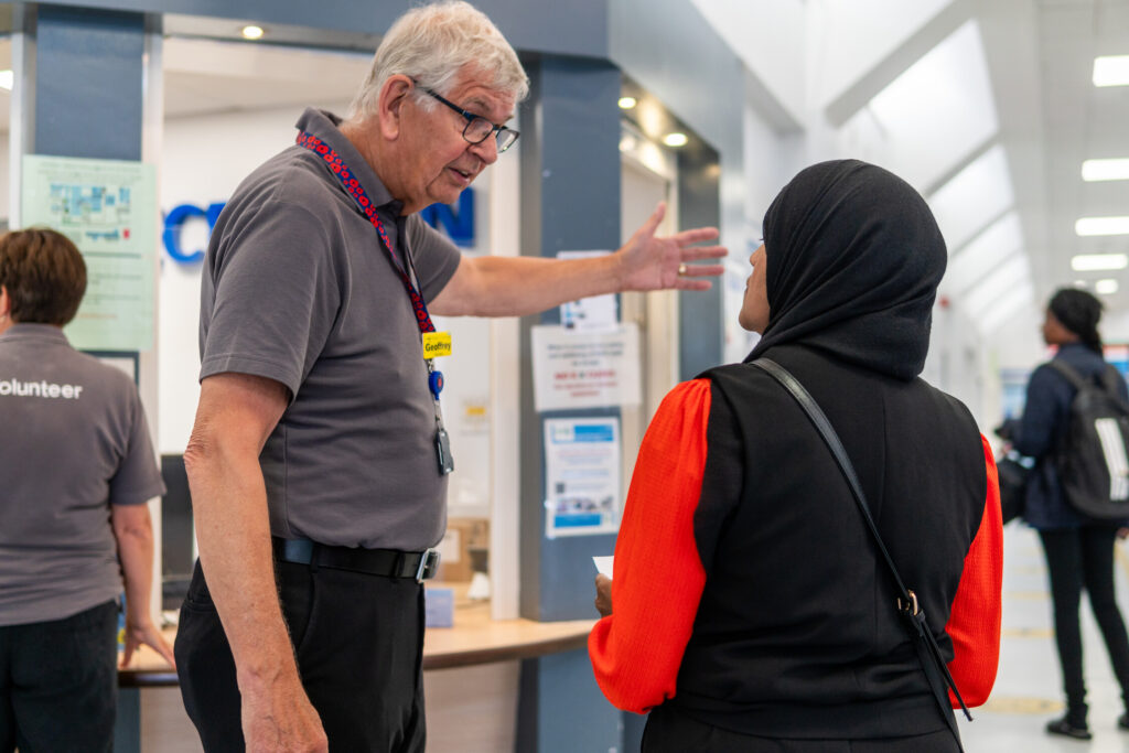 An older man wearing a volunteer badge gestures while speaking to a woman in a black headscarf and red sleeves inside a public building, possibly a hospital or clinic. Other people are visible in the background.