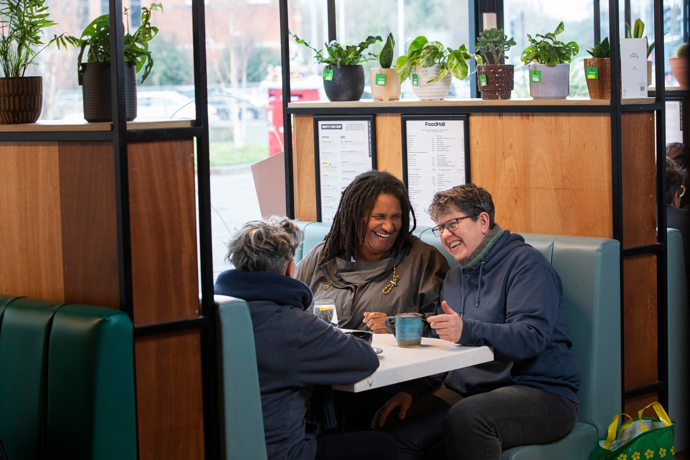 Three people talking at a café table with teal booth seating, potted plants, and large windows in the background.