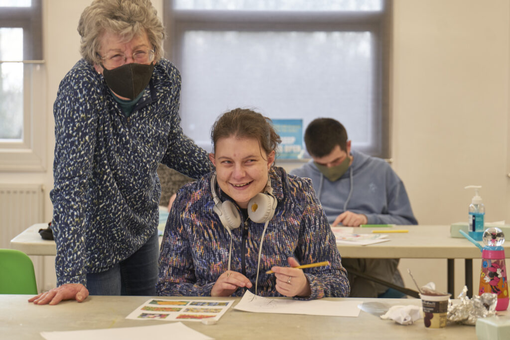 A young woman is sat at a desk writing on a piece of paper and smiling while a member of staff is standing next to her with her hand on the younger person's back.