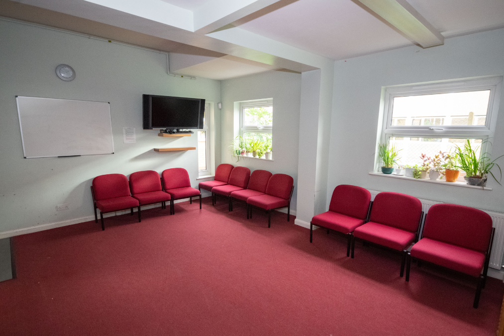Room in Romford Baptist Church with red carpet, red chairs in an L-shape, whiteboard, TV, and potted plants on window sills.