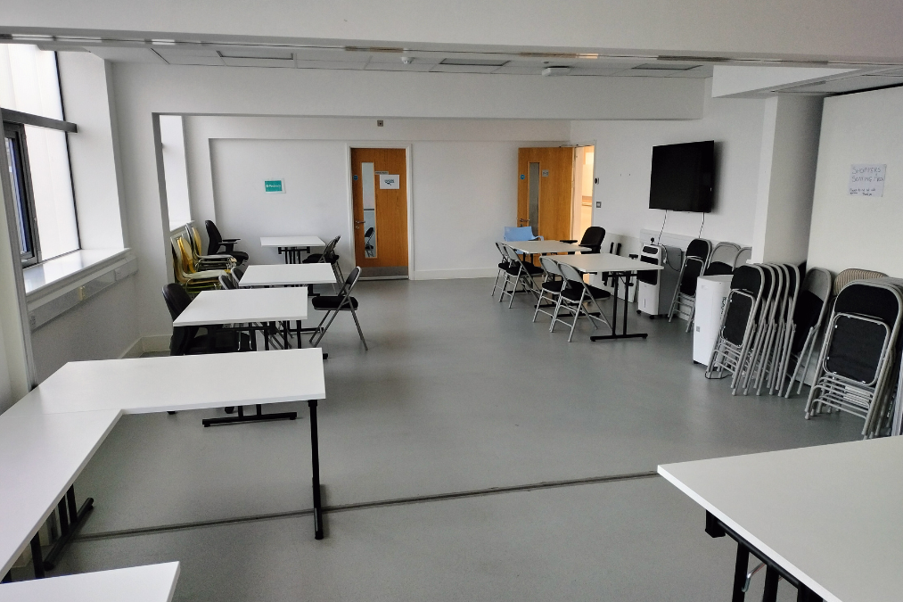 Empty room in Harold Hill library with white tables, chairs, stacked folded chairs, wall-mounted TV, wooden doors, and windows letting in natural light.
