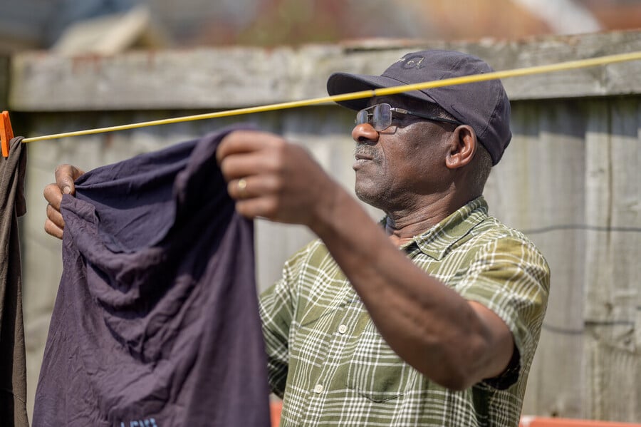 An older man wearing glasses, a hat, and a green plaid shirt hangs laundry on a clothesline outdoors, with a wooden fence in the background.