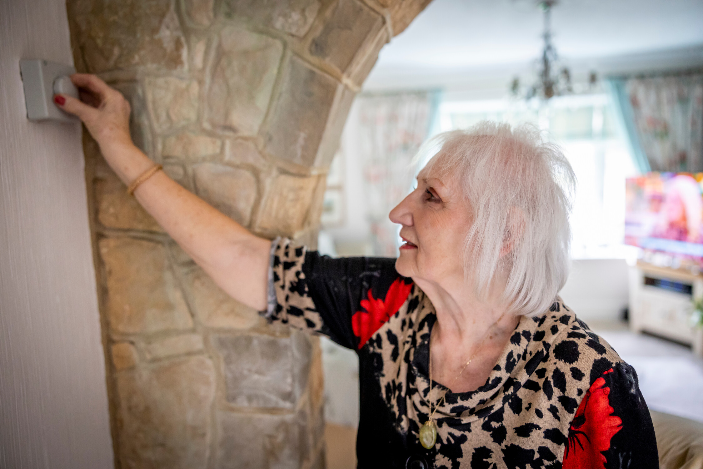 Older woman turning down the temperature on a thermostat on the wall in her house.