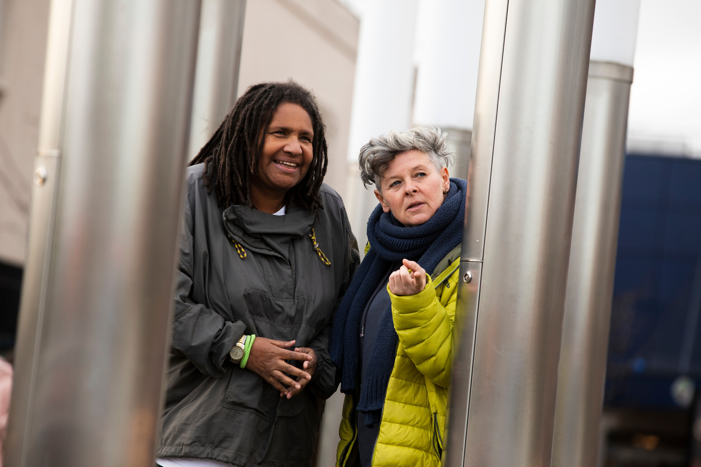 Two women stand outdoors among tall metal pillars. One, with dreadlocks, smiles and wears a gray jacket. The other, with short gray hair and a yellow coat, gestures with her hand while looking forward.