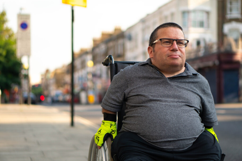 Person using a wheelchair on a city sidewalk, wearing yellow gloves. The background includes buildings and street activity.