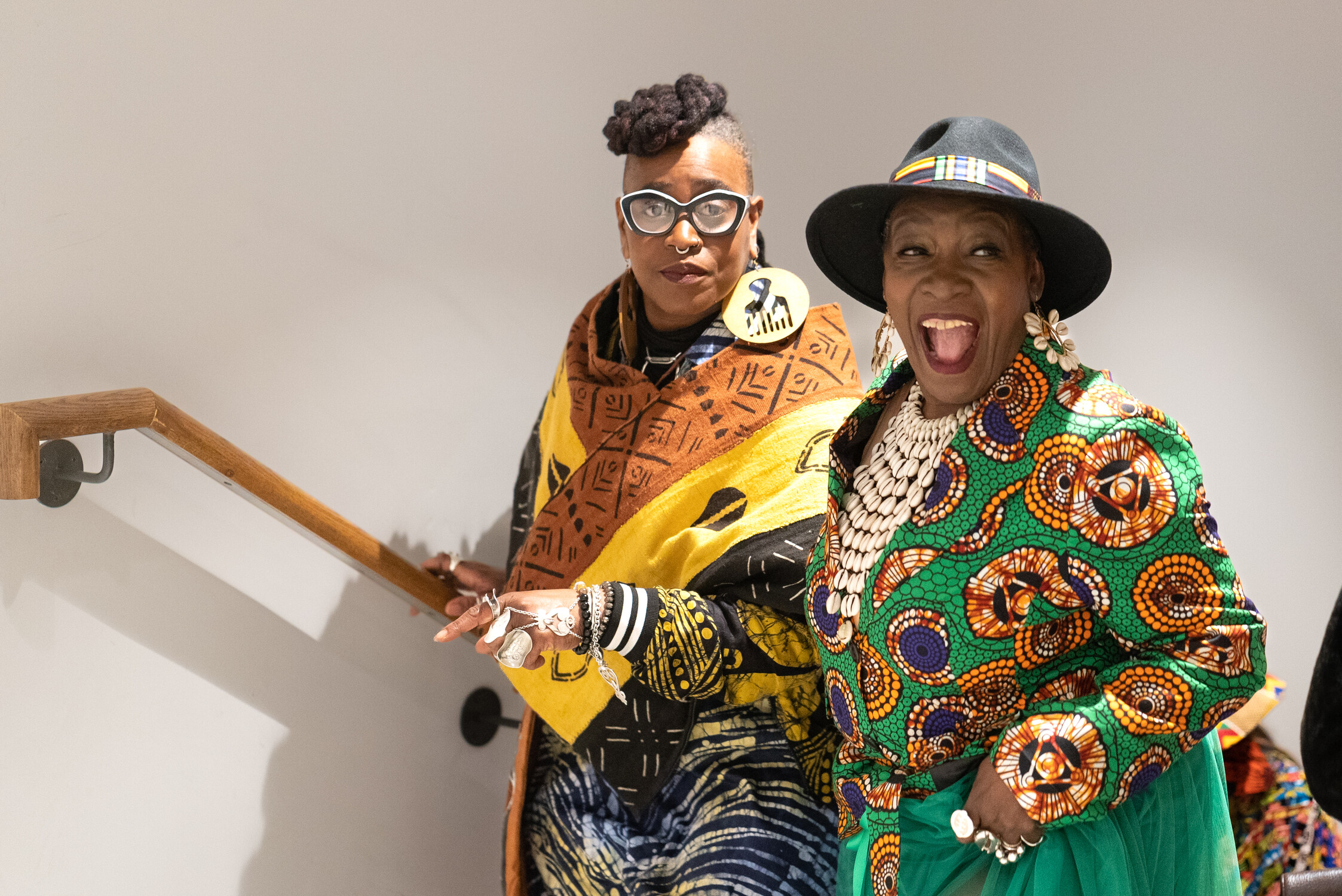 Two women in vibrant, patterned African-inspired clothing and accessories smile on a staircase. One laughs joyfully, while the other stands behind her, both showcasing bold jewelry and unique eyeglasses.