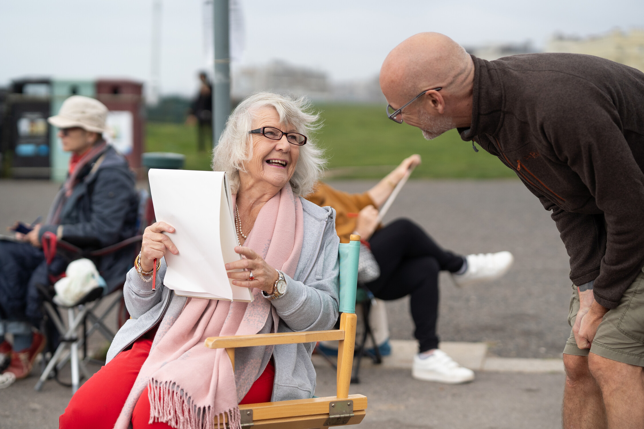 A lady sits on a chair with a sketch pad smiling to a man looking over her shoulder at the art.
