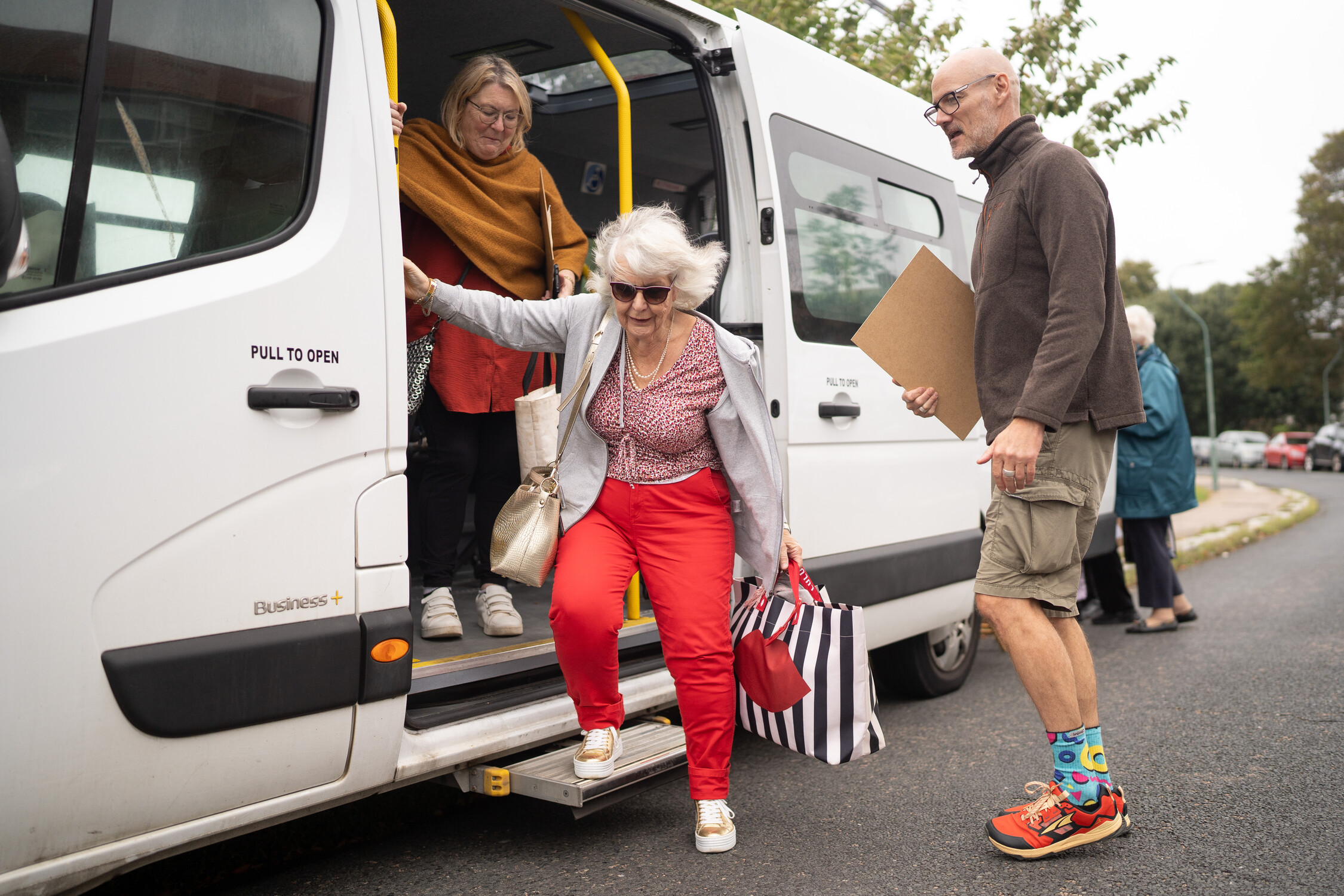 People exiting a white van; one holds a striped bag, another a clipboard.