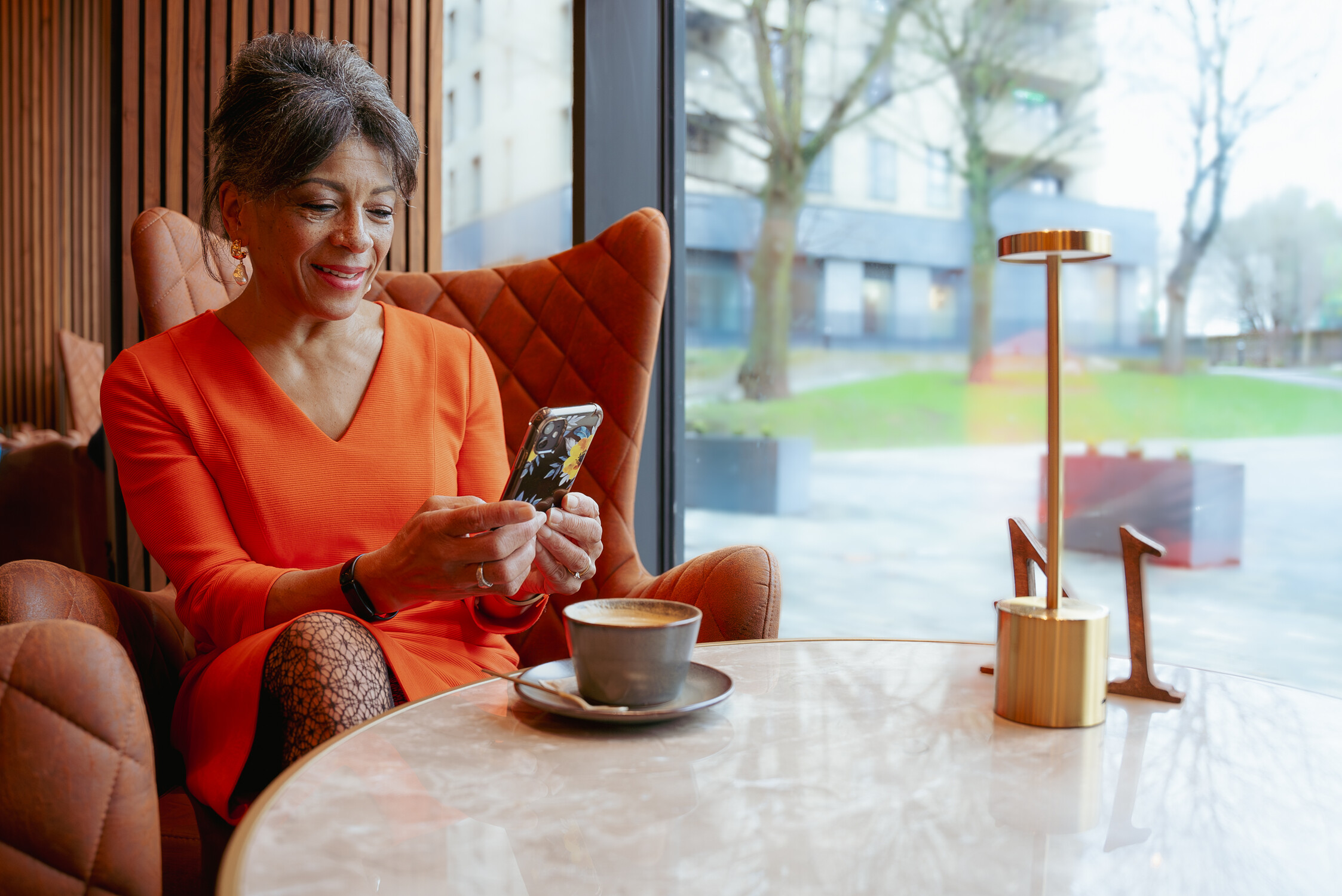 A woman in an orange dress sits in a cozy cafe by a window, smiling as she looks at her smartphone. A cup of coffee and a modern lamp are on the marble table in front of her. Trees and buildings are visible outside.