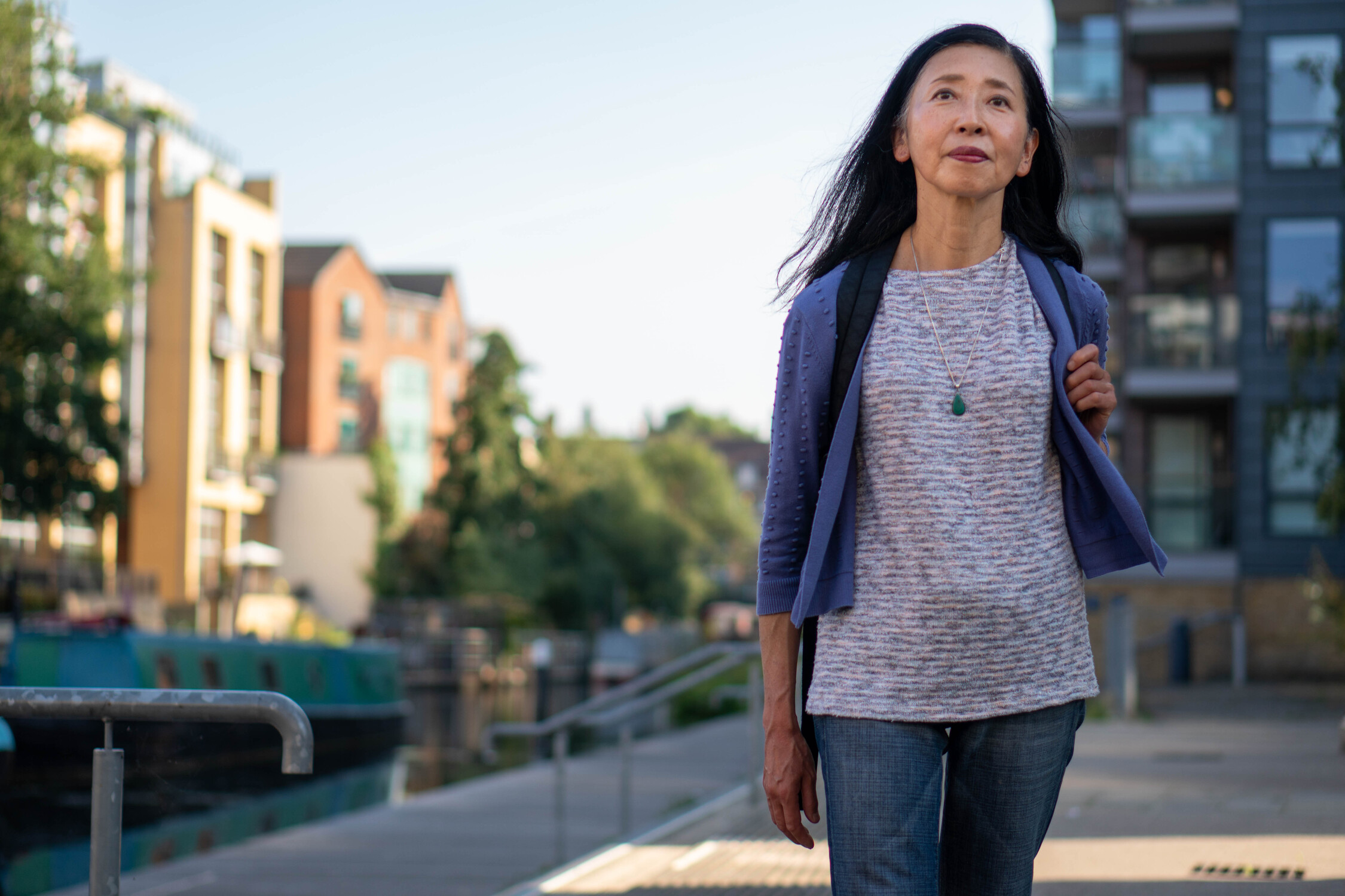 Woman walking near a canal with modern buildings and greenery in the background.