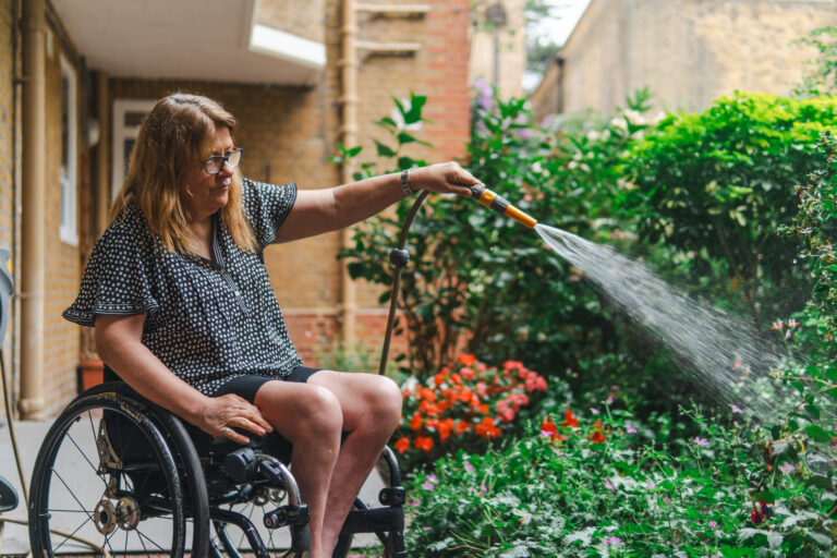 A woman in a wheelchair waters a garden with a hose, surrounded by blooming flowers and green plants on a sunny day.