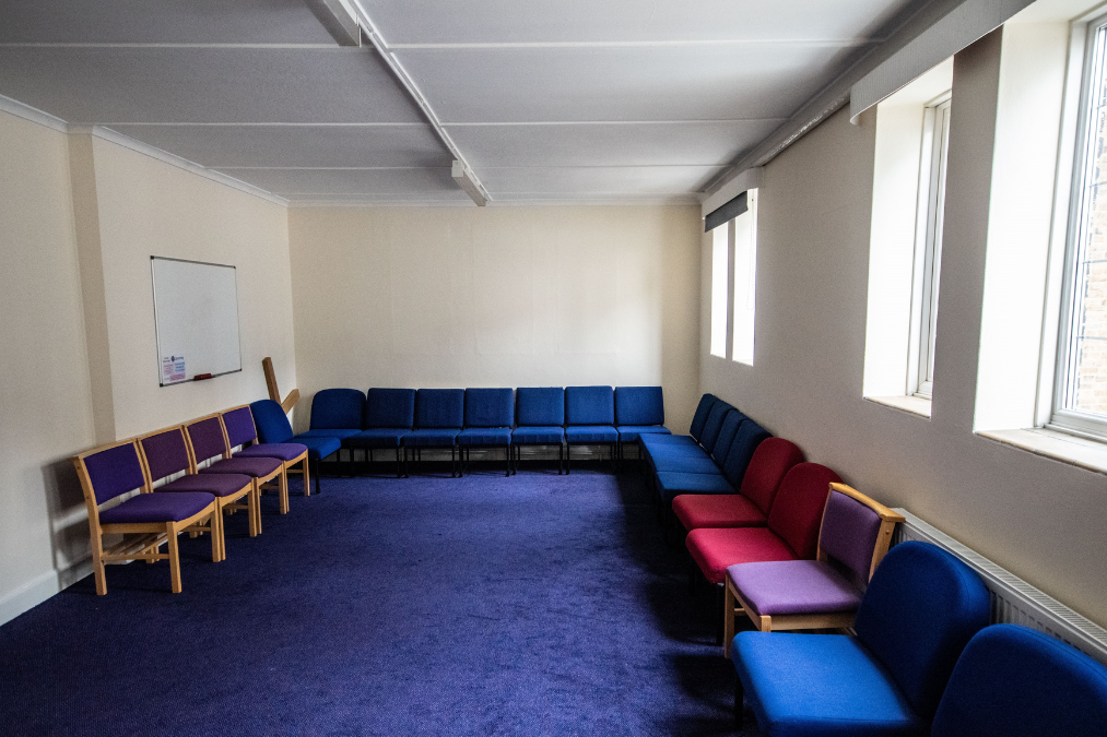 Room in Romford Baptist Church with blue carpet, beige walls, and chairs with colorful cushions arranged in a U-shape; includes a whiteboard and large windows.