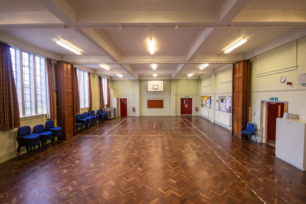 Empty Romford Baptist Church hall with parquet floor, basketball hoop, stacked blue chairs, bulletin boards, and large windows with curtains.