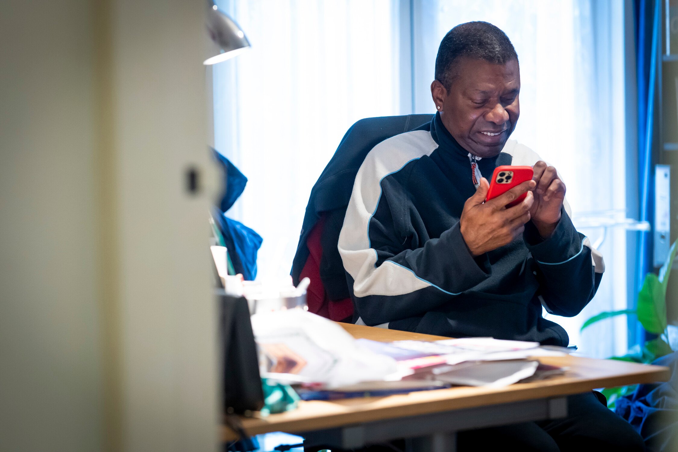 A man sits at a cluttered desk, smiling as he looks at his red phone. He wears a black and white jacket, and the room has bright natural light coming through a window in the background.