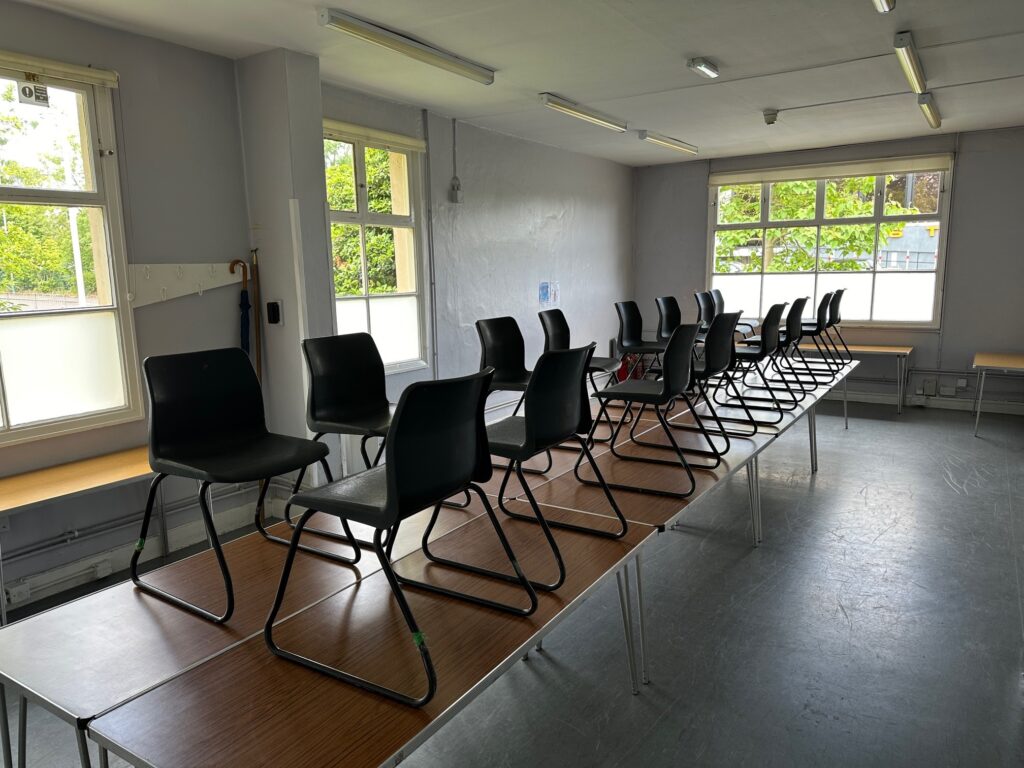 Room at Fairkytes with black chairs on wooden tables, light gray walls, large windows, and ceiling fluorescent lights.
