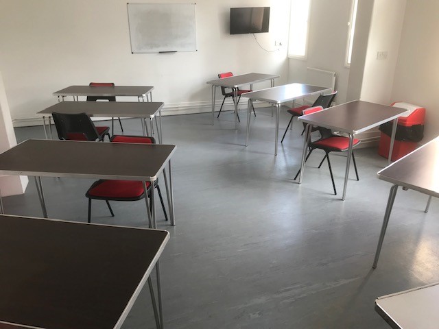 Room in Fairkytes with spaced-out desks and red chairs, a whiteboard and TV at the front, and natural light from windows.