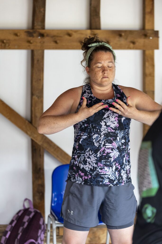 Lady doing yoga in a barn with hands on her chest, wearing a patterned top and grey shorts.