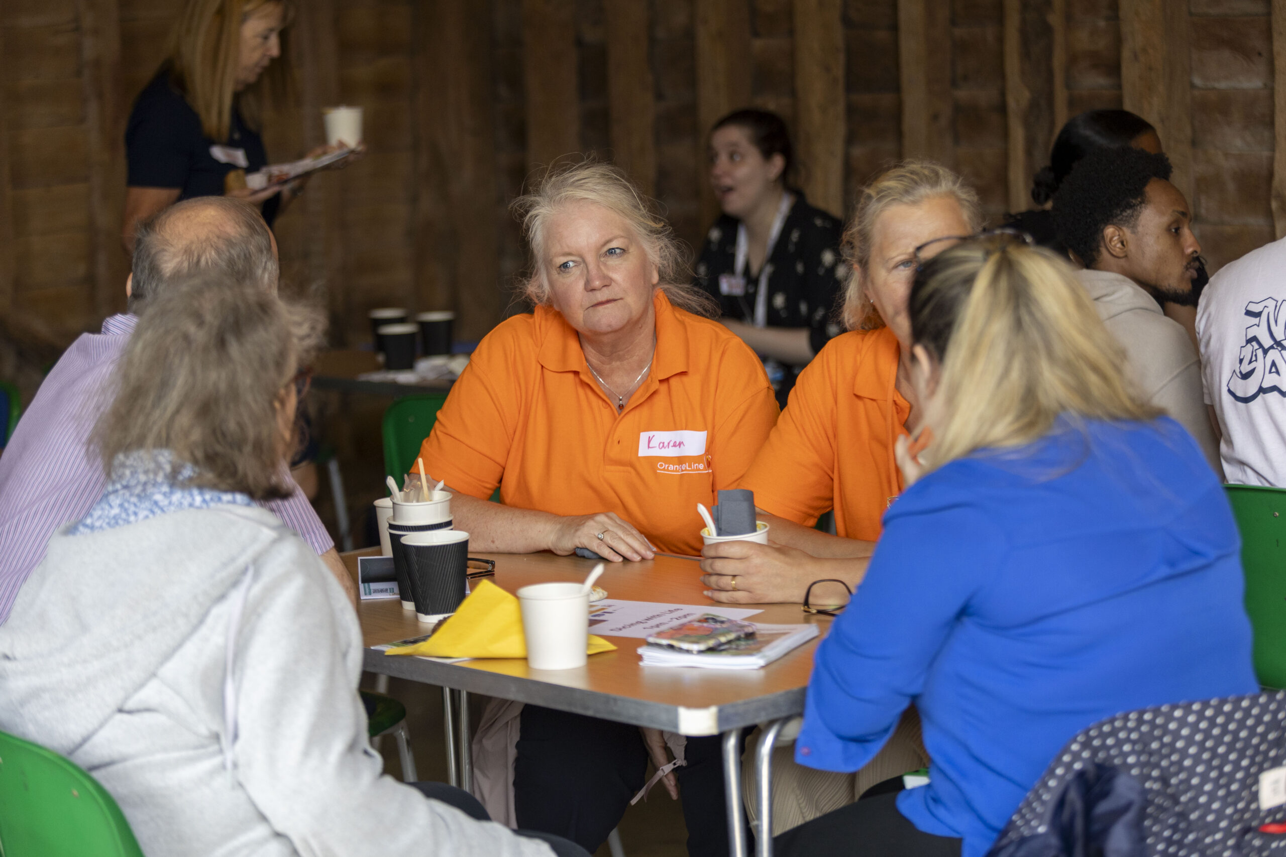 People seated around a table at an indoor gathering, with cups, papers, and a smartphone on the table.